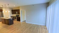 This single-floor space showcases an open-concept design that seamlessly connects the kitchen and living area. The living space features light-toned hardwood flooring that runs throughout, lending warmth and continuity to the interior. Neutral-toned walls in soft gray and warm beige create a bright, welcoming atmosphere that adapts well to various décor styles.

The kitchen is outfitted with dark espresso-toned cabinetry that provides a striking contrast against the lighter walls and flooring. Stainless steel appliances, including a range and over-the-range microwave, are integrated into the cabinetry, contributing to a cohesive and modern aesthetic. Light-toned countertops complete the kitchen's contemporary look.

A doorway visible from the main living area leads to an additional room, suggesting a practical and well-organized floor plan. Multiple electrical outlets are positioned along the walls, offering flexibility for furniture arrangement. Recessed lighting in the kitchen adds focused illumination, while the overall layout prioritizes functionality. The space presents a clean, move-in-ready environment well-suited for everyday living.