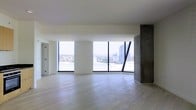 This single-floor space showcases a clean, contemporary design defined by an abundance of natural light and an open layout. The main living area is anchored by large, floor-to-ceiling windows framed in sleek black metal, offering expansive views of the surrounding cityscape, including mid-rise buildings and urban rooftops stretching into the distance. The generous glazing floods the interior with daylight, creating a bright and airy atmosphere throughout.

Light-toned hardwood flooring runs across the entire space, contributing to a sense of warmth while complementing the minimalist aesthetic. A standout architectural feature is the exposed concrete column and wall section positioned toward one side of the room, adding an industrial contrast to the otherwise crisp white walls and ceiling. This raw material detail introduces textural depth and visual interest to the space.

The open-plan layout provides considerable flexibility in how the space can be configured and utilized. The unobstructed sightlines from one end to the other reinforce the spacious feel, making it well-suited for a variety of arrangements. Overall, the space combines modern design sensibilities with urban panoramic views.
