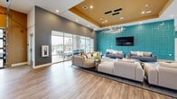 Located in Camby, Indiana, this single-floor community clubhouse is a well-appointed gathering space defined by contemporary design and vibrant, thoughtful interiors. The space is anchored by a bold teal accent wall adorned with geometric paneling, which serves as a striking visual centerpiece throughout. The main lounge area features soaring ceilings with a warm golden-yellow tray detail, illuminated by a modern geometric chandelier and recessed lighting. Multiple seating arrangements with neutral-toned sofas and accent pillows create comfortable, inviting conversation areas. Large floor-to-ceiling sliding glass doors flood the lounge with natural light and provide direct access to an outdoor patio, offering views of the surrounding community. The flooring transitions seamlessly between light wood-look planks and large-format tile, lending the space a clean, cohesive aesthetic. A dedicated kitchen bar area features a generous granite-topped island with modern pendant lighting and stylish barstools, alongside a coffee and refreshment station. The welcoming entry foyer continues the teal accent wall theme, accented with curated artwork and lush potted plants. Throughout the space, the interplay of warm and cool tones, abundant natural light, and modern furnishings creates a polished, community-centered atmosphere.