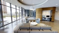 This single-floor space showcases a refined, contemporary design defined by sweeping curved ceilings with recessed lighting and a prominent pendant light as a focal centerpiece. The open-plan layout flows seamlessly between distinct zones, beginning with a generous lounge area furnished with a large sectional sofa in soft grey, paired with rounded accent chairs and a sculptural white bowl coffee table. Geometric decorative objects add an artistic touch to the seating arrangement.

Floor-to-ceiling windows along one wall flood the interior with abundant natural light while offering open views of the surrounding landscape, enhancing the sense of spaciousness throughout. The polished flooring reflects light and contributes to the clean, airy atmosphere. A warm wood-paneled curved partition anchors one side of the space, providing visual contrast against the cool blue-grey tones of the walls. Adjacent to this feature wall is a dedicated workstation area equipped with a computer terminal. A secondary seating area with lighter furnishings and chairs occupies the background near the windows. The overall ambiance is professional yet inviting, blending functional design with modern aesthetic appeal.