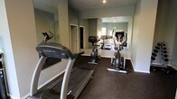 This single-floor fitness room is designed to provide residents or users with a dedicated space for cardiovascular exercise. The room is outfitted with a selection of True-brand cardio equipment, including at least one treadmill, a stationary upright bike, and an elliptical trainer, offering a well-rounded range of low-impact workout options.

The space features durable rubber flooring in a dark tone, which is practical for high-traffic exercise areas and provides both grip and cushioning underfoot. The walls are painted in a soft sage green, creating a calm and focused atmosphere conducive to exercise.

Large wall-mounted mirrors line one side of the room, visually expanding the space and allowing users to monitor their form during workouts. Recessed lighting in the ceiling provides adequate illumination throughout. A window at the far end of the room introduces natural light, enhancing the overall brightness and openness of the space. A door on the side wall provides access to an adjacent area. The overall layout is efficient and functional, making good use of the available floor space.