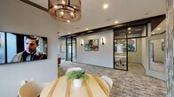 Located at 6515 W Hausman Rd in San Antonio, Texas, this single-story professional office space combines modern design with highly functional workspaces. The interior is defined by a neutral gray color palette, dark crown molding accents, and patterned carpeting that runs throughout, creating a cohesive and polished aesthetic.

The main corridor is wide and well-lit, featuring recessed lighting, wall-mounted artwork, and a warm wood-accented credenza that adds a refined touch to the space. Open seating areas with round tables and ergonomic chairs provide comfortable informal meeting spots, complemented by pendant chandeliers with an industrial-modern style.

A striking textured accent wall in teal and blue tones serves as a bold focal point in one of the open gathering areas, which also benefits from large windows that bring in natural light and views of the surrounding neighborhood.

Glass-walled conference rooms with black metal framing offer both transparency and privacy, each equipped with wall-mounted displays. A dedicated conference room features a long white table with seating for several guests and pendant lighting with an industrial design. A separate breakout nook, finished in deep blue tones with vibrant orange lounge chairs, offers a more intimate and creative setting. Together, these spaces deliver a versatile, design-forward environment suited for professional collaboration.
