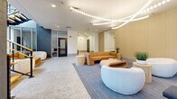 Located at 518 W Fayette St in downtown Baltimore, this single-floor lobby and hallway space offers a well-appointed entry experience spanning a generous footprint. The design blends contemporary aesthetics with functional circulation, creating an inviting first impression for occupants and visitors alike.

The lounge area serves as the centerpiece of the lobby, featuring terrazzo flooring that transitions into a carpeted seating zone. Furnishings include a plush mustard-toned sofa, curved cream accent chairs, and cylindrical side tables, all anchored beneath a striking serpentine pendant light installation that traces an elegant path across the ceiling. Floor-to-ceiling windows flood the space with natural light and offer views of the surrounding streetscape.

A sculptural open staircase with warm wood treads and brass railings rises prominently from the lounge, framed by a dark tiled accent wall and additional glazed facades that reinforce the sense of openness and vertical connection.

The elevator corridor extends from the lounge, finished with vertical wood slat wall paneling, polished stainless steel elevator doors, and a dark drop ceiling with recessed lighting. Terrazzo flooring continues throughout, tying the spaces together cohesively. A secondary access door with porthole windows completes the functional hallway layout, ensuring smooth building circulation throughout.