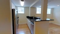 This single-floor space showcases a thoughtfully designed kitchen that serves as its centerpiece. The kitchen features a galley-style layout with sleek dark granite countertops that create a striking contrast against the crisp white cabinetry below. Stainless steel appliances, including a full-size refrigerator and dishwasher, add a modern, polished touch to the space. A peninsula with matching dark granite extends from the counter, offering additional workspace or casual seating. Warm-toned hardwood flooring runs throughout, lending a sense of continuity and natural warmth to the interior. The walls are finished in a soft, neutral tone that keeps the space feeling bright and welcoming. Two windows allow natural light to filter in, illuminating the kitchen and providing views of the surrounding neighborhood. Overhead lighting ensures the space remains well-lit at all times. The open layout and clean design create an inviting, functional environment that balances practicality with contemporary style, making it a comfortable and efficient space for everyday living.