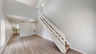 This two-story loft in Baton Rouge, Louisiana offers a smart and functional layout that maximizes vertical space through dramatic vaulted ceilings and an open-concept design. The main level features a spacious living area with soaring ceilings, light gray walls, and durable wood-style flooring that flows throughout. Large windows allow natural light to fill the space, creating a bright and airy atmosphere. A white-railed staircase serves as a central architectural feature, connecting the main level to the upper loft. The kitchen is equipped with white cabinetry, a coil electric range, a full-size refrigerator, a stainless steel sink, and a window overlooking the outdoors, providing natural light while cooking. A convenient half bathroom is located on the main level, fitted with a single vanity, speckled countertop, and mirror. Upstairs, the loft-style bedroom features sloped ceilings, warm wood-tone flooring, and multiple windows that bring in additional natural light. The upper level includes a walk-in closet and a full bathroom with a matching vanity and countertop. The open loft railing offers a sense of connectivity between floors, enhancing the overall open and airy character of this well-designed Baton Rouge residence.
