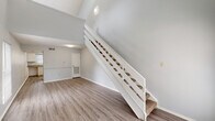 This two-story loft in Baton Rouge, Louisiana offers a smart and functional layout that maximizes vertical space through dramatic vaulted ceilings and an open-concept design. The main level features a spacious living area with soaring ceilings, light gray walls, and durable wood-style flooring that flows throughout. Large windows allow natural light to fill the space, creating a bright and airy atmosphere. A white-railed staircase serves as a central architectural feature, connecting the main level to the upper loft. The kitchen is equipped with white cabinetry, a coil electric range, a full-size refrigerator, a stainless steel sink, and a window overlooking the outdoors, providing natural light while cooking. A convenient half bathroom is located on the main level, fitted with a single vanity, speckled countertop, and mirror. Upstairs, the loft-style bedroom features sloped ceilings, warm wood-tone flooring, and multiple windows that bring in additional natural light. The upper level includes a walk-in closet and a full bathroom with a matching vanity and countertop. The open loft railing offers a sense of connectivity between floors, enhancing the overall open and airy character of this well-designed Baton Rouge residence.