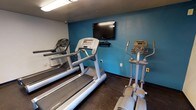 This single-floor fitness center offers a focused and functional workout environment designed to meet essential cardio training needs. The room features a bold teal accent wall that adds a vibrant, energetic atmosphere, complemented by neutral grey walls and durable dark rubberized flooring ideal for high-traffic exercise areas. The space is equipped with multiple treadmills and elliptical cross-trainers, providing a variety of cardio options for users. A wall-mounted flat-screen television is positioned at eye level, allowing guests to stay entertained during their workouts. Overhead fluorescent lighting ensures the room is well-lit throughout. The layout is straightforward and efficient, maximizing the use of available floor space while keeping equipment accessible and easy to navigate. The overall ambiance is practical and purposeful, catering to those looking for a convenient and no-frills fitness facility.