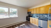 This well-appointed 1-bedroom, 1-bathroom unit at 510 Maloney Ave in Poughkeepsie, NY offers a comfortable and functional single-floor layout. The kitchen is a standout feature, equipped with stainless steel appliances including a range, microwave, refrigerator, and dishwasher. Warm-toned wood cabinetry pairs with a stylish mosaic tile backsplash and dark countertops, while hardwood-style flooring runs throughout the space. A window above the kitchen area allows natural light to flood in, offering views of the surrounding greenery. Adjacent to the kitchen, a dining or flex area provides additional living space, also finished with hardwood-style flooring. The spacious living room is carpeted and features a large picture window that frames lush outdoor views, along with a baseboard heating system and a wall-mounted air conditioning unit for year-round comfort. The bedroom is generously sized with soft carpeting, neutral walls, and access to a closet. The full bathroom is clean and bright, featuring tile flooring, a white vanity, and a tub/shower combo with a window for natural ventilation. A decorative wrought-iron stair railing is also visible, adding a subtle architectural detail to the space.