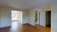 This two-story home presents a welcoming and straightforward layout designed for comfortable everyday living. The main level features warm hardwood flooring that flows seamlessly through the open living areas, lending a cohesive and inviting feel to the space. A wide interior archway connects the main living room to an adjacent room, which is bathed in natural light from a large window overlooking a lush, green outdoor view. This connected layout creates a generous and airy sense of space, ideal for both relaxing and entertaining. Baseboard heating runs along the perimeter of the lower level, ensuring year-round comfort. A dedicated staircase provides access to the upper floor, positioned alongside a classic paneled door that adds a traditional architectural touch. The walls throughout are finished in a soft, neutral tone that complements the natural warmth of the flooring and keeps the interior feeling bright and clean. The overall ambiance of the home is calm and understated, with a practical design that emphasizes functionality. The two-floor configuration offers a natural separation between living and private spaces, making this home a well-organized and versatile residence.