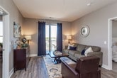 This single-floor residence showcases a cohesive contemporary design carried consistently through each room. The kitchen serves as a functional centerpiece, featuring rich dark cabinetry contrasted against light granite countertops and a classic white subway tile backsplash. Stainless steel appliances, including a range, microwave, dishwasher, and refrigerator, provide a polished, modern aesthetic, while a built-in wine rack adds a distinctive design detail. Wood-style flooring grounds the space with warmth and durability.

The living area offers a welcoming atmosphere, highlighted by large windows and sliding glass door access to a private balcony, allowing natural light and outdoor views to enhance the interior ambiance. Neutral gray walls provide a versatile backdrop throughout the unit.

The bedroom is carpeted for comfort and includes a ceiling fan, natural light from a generously sized window, and ample closet access. The room comfortably accommodates a queen-sized bed with space for additional furnishings.

Two bathrooms serve the residence, each finished with matching dark cabinetry, granite countertops, and wood-style flooring. One bathroom features a shower-tub combination, while the second offers a walk-in shower and open shelving for storage, completing a well-designed and functional living space.