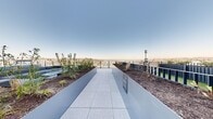 Located at 757 Thomas St in Seattle's Celestial Terrace neighborhood, this striking rooftop terrace sits atop a modern high-rise clad in sleek architectural paneling and floor-to-ceiling glass. The outdoor space wraps around the building, offering sweeping 360-degree views of the Seattle skyline, Elliott Bay, and the city's surrounding residential and commercial districts.

The terrace is thoughtfully divided into multiple functional zones. A wide, light-toned paver walkway runs along the perimeter, bordered by glass panel railings that preserve unobstructed sightlines to the cityscape below. Raised planters filled with ornamental grasses and native plantings add a green, living element throughout. A dedicated outdoor kitchen area features a built-in stainless steel grill and bold blue cabinetry, ideal for entertaining. Nearby, a spa or hot tub area with a tiled surround, wooden deck, and lounge chairs provides a relaxing retreat with direct views of downtown Seattle. Additional seating areas with bistro tables and woven chairs are positioned along the terrace edges, maximizing exposure to natural light and the dramatic urban panorama. The overall design blends contemporary architecture with thoughtful landscaping, creating a refined and versatile elevated outdoor environment.