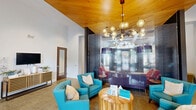 Located on McQueen Drive in Durham, NC, this single-floor community space offers a thoughtfully designed interior that balances function with contemporary style. The grand entrance greets visitors with soaring ceilings, an arched transom window, and floor-to-ceiling glass doors that flood the lobby with natural light. Elegant sheer drapery, teal upholstered seating, and a modern sputnik-style chandelier set a welcoming tone from the moment you step inside.

The open layout flows seamlessly into multiple lounge areas, each with its own distinct character. One section features a warm wood-paneled ceiling that creates an intimate atmosphere, complemented by vibrant turquoise armchairs and a textured glass accent wall. Another area showcases a coffered ceiling with recessed and pendant lighting, bold colorful artwork, and an eclectic mix of seating including woven accent chairs, red wingbacks, and a plush sectional — ideal for both casual gatherings and structured meetings.

Throughout the space, durable neutral-toned flooring ties the zones together, while carefully curated furnishings and accent pieces add pops of color and personality. Private rooms with glass-panel doors provide quieter breakout spaces, making this a highly versatile and inviting community hub in the heart of Durham.