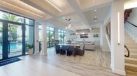 Located at 451 Hialeah Dr in Hialeah, Florida, this contemporary two-story amenity space is designed with a clean, modern aesthetic that emphasizes open layouts, natural light, and sophisticated finishes. Light wood-look flooring flows consistently throughout both levels, complemented by crisp white walls and coffered ceiling details that add architectural depth.

The main level entry presents a bright, expansive lobby with large floor-to-ceiling glass doors and windows that frame views of lush tropical palms and the surrounding outdoor areas. A long bar-height counter with blue upholstered stools anchors one side of the entry, while pendant lighting in varying styles adds warmth and visual interest overhead.

Multiple distinct gathering areas are distributed across both floors. A well-appointed kitchen features light wood cabinetry reaching to the ceiling, a generous white waterfall island with blue barstools, and gold pendant fixtures. A second kitchen area offers bold navy cabinetry, an island with leather-toned stools, and large windows overlooking greenery.

Lounge areas are furnished with plush neutral sectionals, statement chandeliers, wall-mounted televisions, and built-in cabinetry. A dedicated dining room showcases a long communal table, sculptural wired pendant lights, and a decorative geometric partition wall. A staircase connects both floors, with glass-paneled doors and corridor seating completing the thoughtfully designed layout.
