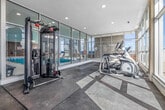 Located at 225 Davisville Ave in Toronto's Davisville Village neighbourhood, this fitness centre offers a well-equipped, single-floor workout space designed with both functionality and comfort in mind. The open-concept layout is bathed in natural light, courtesy of expansive floor-to-ceiling windows that line multiple walls and provide views of the surrounding rooftop and cityscape.

The space is finished with a clean, modern aesthetic throughout — white walls, recessed lighting, and interlocking foam and hard flooring panels that define workout zones. The strength training area features a multi-station cable machine, a tiered dumbbell rack with a full range of free weights, a adjustable weight bench, and exercise mats for stretching or floor work. A large wall-mounted mirror enhances the sense of space and supports training form.

The cardio zone, situated along the window-lined perimeter, houses elliptical trainers and a stationary bike, taking full advantage of the natural light and open sightlines. Exposed brick accents near the entry corridor add a touch of architectural character, complementing the otherwise contemporary design. A hand sanitizing station and waste disposal are conveniently positioned near the entrance. The fitness centre is also adjacent to a pool area, visible through interior glass partitions, offering a well-rounded amenity experience.