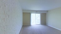 This single-floor space presents a clean, open layout with a notably bright ambiance. One of the most distinctive architectural features is a full textured brick-style accent wall that adds visual depth and character to the room. The neutral beige walls complement the light-toned carpeted flooring, creating a cohesive and understated aesthetic throughout.

A central highlight of the living area is the sliding glass door, dressed with vertical blinds, which opens onto a balcony with a wrought-iron railing. This feature allows ample natural light to fill the interior while providing a view of surrounding greenery, contributing to an airy and inviting atmosphere. The overhead HVAC vent integrated into the ceiling soffit indicates a central air system, supporting year-round comfort.

The space is currently unfurnished, allowing for a clear appreciation of its layout and proportions. The combination of the textured accent wall, soft carpeting, and generous natural light from the sliding door creates a balanced environment suitable for a comfortable living area. The balcony access further enhances the connection between the interior and the outdoors.