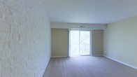 This single-floor space presents a clean, open layout with a notably bright ambiance. One of the most distinctive architectural features is a full textured brick-style accent wall that adds visual depth and character to the room. The neutral beige walls complement the light-toned carpeted flooring, creating a cohesive and understated aesthetic throughout.

A central highlight of the living area is the sliding glass door, dressed with vertical blinds, which opens onto a balcony with a wrought-iron railing. This feature allows ample natural light to fill the interior while providing a view of surrounding greenery, contributing to an airy and inviting atmosphere. The overhead HVAC vent integrated into the ceiling soffit indicates a central air system, supporting year-round comfort.

The space is currently unfurnished, allowing for a clear appreciation of its layout and proportions. The combination of the textured accent wall, soft carpeting, and generous natural light from the sliding door creates a balanced environment suitable for a comfortable living area. The balcony access further enhances the connection between the interior and the outdoors.