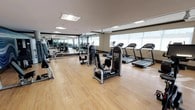This fitness center offers a thoughtfully designed workout space that balances functionality with a clean, modern aesthetic. The room features warm-toned hardwood flooring throughout, providing a polished and durable surface suited for a variety of exercise activities. Large floor-to-ceiling windows line one side of the space, flooding the interior with generous natural light and creating an open, airy atmosphere that enhances the overall workout experience.

Full-length mirrored walls run along the opposite side, visually expanding the room and allowing users to monitor their form during exercise. Recessed and flush-mounted ceiling lights supplement the natural light, ensuring the space remains well-illuminated at all times.

The fitness center is equipped with a comprehensive selection of machines, including multiple treadmills positioned near the windows to take advantage of natural light, as well as a range of strength-training cable and resistance machines arranged across the floor. The layout is organized and spacious, with ample room between equipment for safe and comfortable movement. The neutral color palette of grays, blacks, and warm wood tones gives the space a contemporary, professional feel suited for focused, effective training sessions.
