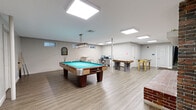 Located at 90 Gas Light Dr in Weymouth, Massachusetts, this lower-level community amenity space offers a well-appointed recreational area designed for entertainment and relaxation. The open-plan layout is anchored by a full-size pool table positioned beneath a stylish pendant light fixture, creating a natural focal point throughout the room. Light wood-look flooring runs throughout, complementing the neutral gray walls and bright flush-mount panel lighting that keeps the space feeling open and well-illuminated.

A striking exposed brick fireplace with a white mantel serves as a standout architectural feature, adding warmth and character to the room. Alongside the pool table, the space accommodates an air hockey table and a foosball table, providing a variety of recreational options. A dedicated computer workstation with a desk and chair offers a practical workspace within the same area.

An adjacent fitness room is visible through a connecting doorway, equipped with cardio machines for residents seeking exercise options. The space is accessed via a carpeted staircase and features clearly marked exits, ensuring safety and convenience. Small windows near the ceiling allow for modest natural light, while the overall design balances functionality with a comfortable, community-oriented atmosphere.