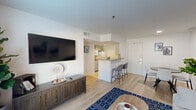Located at 7880 Alpha Rd in Dallas, TX, this well-appointed one-bedroom, two-bathroom residence offers a thoughtfully designed single-floor layout. Light wood flooring flows throughout the open-concept living area, creating a cohesive and airy feel. The living room is bathed in natural light through large windows dressed with teal drapes, offering views of the lush outdoor greenery. White walls and smooth ceilings complement the clean, modern aesthetic.

The kitchen features light cabinetry, granite-style countertops, and a peninsula breakfast bar with seating — seamlessly connecting to the dining and living areas for easy entertaining. A dedicated dining space provides room for a full table setup alongside the main living area.

The bedroom is a comfortable retreat, complete with a ceiling fan, a large window with natural light, and direct access to a full bathroom. The en-suite bath features white tile flooring, an extended vanity with an integrated sink, and a walk-in closet with built-in storage. A second full bathroom serves guests. Throughout the residence, the design prioritizes functionality and a clean, contemporary style suitable for everyday living in a prime Dallas location.