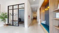 Located at 2703 Girard Ave S in Minneapolis, MN, the NOX building presents a thoughtfully designed lobby and hallway that serves as a welcoming first impression for residents and visitors alike. The single-floor space features polished concrete floors that reflect abundant natural light pouring in through expansive floor-to-ceiling windows at the building entrance, offering clear views of the surrounding Uptown neighborhood streetscape.

The layout includes a glass-enclosed leasing office positioned prominently off the main hallway, framed by sleek black steel partition walls that lend a contemporary, industrial-chic aesthetic. Inside the leasing office, soft blue-gray walls, carpeted flooring, and casual seating create an approachable and professional environment.

A standout architectural feature is the custom built-in seating nook, finished in warm wood tones with a teal upholstered bench and dark textured backsplash illuminated by amber accent lighting. Flanking this nook is an open wood shelving unit displaying decorative objects, adding character and warmth to the space.

The main hallway extends deeper into the building with clean white walls, recessed ceiling lighting, and a mailbox bank, providing residents with convenient access to essential services. The overall ambiance balances modern minimalism with warm, inviting design elements, creating a polished and functional communal entrance.