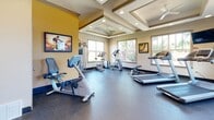 The fitness center at Westbrook Crossing in Collierville, Tennessee, is a thoughtfully designed single-floor space that provides residents with a well-equipped environment for cardiovascular and strength training. The room features a distinctive coffered ceiling with recessed lighting and a ceiling fan, lending the space an architectural elegance uncommon in community fitness facilities. Warm neutral and yellow accent walls contribute to an energizing atmosphere, while motivational artwork and framed cycling-themed pieces reinforce the active ambiance throughout.

Multiple windows line the walls, allowing generous natural light to fill the room and offering views of the surrounding greenery, creating an inviting and open feel. The durable rubberized flooring provides a safe and functional surface suited for a range of exercises. Equipment includes treadmills, elliptical machines, upright and recumbent stationary bikes, and a weight bench, offering a comprehensive selection for varied fitness routines. A wall-mounted flat-screen television adds convenience for entertainment during workouts. The layout is spacious enough to accommodate multiple users comfortably, with clear circulation paths between machines. Overall, the fitness center combines practical functionality with attractive design details.