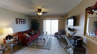 This single-floor space centers around a well-appointed living room that balances functionality with comfort. The room is finished with neutral-toned walls and carpeted flooring, creating a warm and inviting atmosphere. A ceiling fan with an integrated light fixture is mounted overhead, providing both air circulation and ambient lighting.

Natural light flows generously into the space through a large sliding glass door, which opens onto a private balcony with a railing, offering outdoor access and views beyond. The sliding door serves as the room's primary focal point, drawing the eye toward the exterior and enhancing the sense of openness.

The living area is furnished with a plush, deep-red sectional sofa adorned with decorative pillows, paired with a cushioned accent chair. A glass-top coffee table and a matching side console table anchor the seating arrangement, which is grounded by a patterned area rug. A wall-mounted flat-screen television is positioned on the opposite wall alongside an entertainment console, completing the media setup. A tall indoor plant adds a natural, decorative element to the corner of the room, contributing to the overall lived-in and welcoming character of the space.