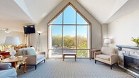 Located at 9950 Redwood St NW in Coon Rapids, Minnesota, this single-floor unit offers a thoughtfully organized layout comprising a living room, a combined kitchen and dining area, and one bedroom. The standout architectural feature is the living room's soaring vaulted ceiling that rises to a dramatic gable peak, framed by a large floor-to-ceiling multi-pane window that floods the space with natural light and provides sweeping views of surrounding mature trees. A stone-surround fireplace anchors one corner of the living room, adding warmth and character to the space. Soft blue-toned carpeting runs throughout the living area, complementing the bright, neutral wall tones. The kitchen features warm oak cabinetry, light countertops, a peninsula with a breakfast bar, and full-size appliances including a refrigerator and dishwasher. Adjacent to the kitchen, the dining area accommodates multiple round tables with Windsor-style chairs, making it well-suited for shared or communal dining. A ceiling fan provides additional ventilation in the kitchen-dining zone. The bedroom is a compact, private retreat. Overall, the space offers a comfortable and functional environment with a bright, open ambiance defined by its signature vaulted living room and generous natural light.
