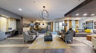 Located at 1700 W Frankford Rd in Carrollton, TX, this thoughtfully designed community clubhouse offers a generous, open-concept floor plan that seamlessly integrates multiple functional areas into one cohesive, inviting space. The interior design is anchored by a striking color palette of charcoal gray, warm gold, and rich wood tones, creating a contemporary yet welcoming ambiance throughout.

The central lounge area serves as the heart of the space, featuring an arrangement of gray upholstered armchairs and sofas surrounding a dark leather cocktail ottoman, all set atop a bold gold-patterned area rug. A sculptural orb chandelier hangs overhead, complementing the recessed lighting and varied ceiling details, including a distinctive wood-paneled dropped ceiling section that adds architectural depth.

Adjacent to the lounge, a kitchen and bar area is outfitted with dark cabinetry, stainless steel appliances, a marble-topped counter, and vivid yellow bar stools beneath sleek pendant lights. A dedicated business center provides workstations set against a dramatic accent wall with abstract artwork. The space also features a media lounge with a mounted flat-screen TV and curated décor displays. Glass-paneled doors lead directly to a fitness center, visible from the main floor. Large windows and glass doors fill the space with natural light.