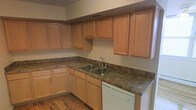 This single-floor space offers a thoughtfully designed layout centered around a functional and inviting kitchen. The kitchen is outfitted with warm-toned natural wood cabinetry that extends to the upper walls, providing generous storage across both upper and lower cabinets. The countertops feature a dark granite-look surface that contrasts beautifully with the lighter wood tones, creating a visually balanced aesthetic. An L-shaped countertop configuration maximizes workspace, making the kitchen both practical and efficient. A stainless steel double-basin sink is positioned along the counter and paired with a sleek chrome faucet, while a white dishwasher is seamlessly integrated beneath the countertop for added convenience. The flooring throughout the kitchen is composed of warm hardwood planks, adding a sense of continuity and natural texture to the space. Neutral-toned walls complement the overall palette, keeping the atmosphere open and clean. The kitchen benefits from a ceiling vent for climate control, ensuring year-round comfort. Overall, the space presents a cohesive, move-in ready environment with classic finishes and practical design elements suited for everyday living.