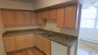 This single-floor space offers a thoughtfully designed layout centered around a functional and inviting kitchen. The kitchen is outfitted with warm-toned natural wood cabinetry that extends to the upper walls, providing generous storage across both upper and lower cabinets. The countertops feature a dark granite-look surface that contrasts beautifully with the lighter wood tones, creating a visually balanced aesthetic. An L-shaped countertop configuration maximizes workspace, making the kitchen both practical and efficient. A stainless steel double-basin sink is positioned along the counter and paired with a sleek chrome faucet, while a white dishwasher is seamlessly integrated beneath the countertop for added convenience. The flooring throughout the kitchen is composed of warm hardwood planks, adding a sense of continuity and natural texture to the space. Neutral-toned walls complement the overall palette, keeping the atmosphere open and clean. The kitchen benefits from a ceiling vent for climate control, ensuring year-round comfort. Overall, the space presents a cohesive, move-in ready environment with classic finishes and practical design elements suited for everyday living.