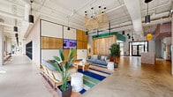 Located at 440 W State St in Columbus, Ohio, this thoughtfully designed single-floor amenity space offers a generous, open-plan layout that blends modern industrial aesthetics with warm, natural materials. Polished concrete floors run throughout, complemented by soaring ceilings with exposed ductwork and an array of woven rattan pendant lights that add texture and warmth to the environment.

The central lounge area serves as the heart of the space, featuring rattan-framed sofas, a live-edge wood coffee table, and a vibrant striped area rug that anchors the seating arrangement. Lush tropical plants are strategically placed throughout, adding a fresh, organic touch. A feature wall with wood paneling and a mounted display screen enhances the communal atmosphere.

Adjacent to the lounge, a bold glass-partitioned fitness center is fully visible, housing treadmills, stationary bikes, and strength equipment, all bathed in natural light from large street-facing windows. A café-style nook with light wood half-walls, hanging greenery, and blush pink bistro tables and chairs provides an intimate space for casual gatherings.

A terrazzo-topped island with reclaimed wood accents serves as a welcoming reception or refreshment station, flanked by vending machines and a utility sink. Accent walls in teal, sage green, and mauve add bold pops of color, while curated artwork and decorative mirrors complete the polished, community-forward ambiance.