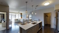 This single-floor space showcases an open-concept layout that integrates the kitchen and living area into one cohesive, functional environment. The kitchen is anchored by a large central island with a crisp white countertop, an undermount double sink, and a built-in dishwasher. Dark wood cabinetry on the island provides a striking contrast to the light surfaces, while three stylish pendant lights hang above, offering both task lighting and a decorative focal point. The island also serves as a casual dining space, with seating and place settings visible along one side.

The living area flows naturally from the kitchen, featuring a light-toned sofa accented with decorative pillows in teal and yellow. A ceiling fan overhead and large windows dressed with teal and gold curtains allow natural light to fill the space. Dark hardwood-style flooring runs throughout, grounding the open layout with warmth and continuity. A glimpse into an adjacent room reveals a similarly styled bedroom. The overall ambiance is modern and well-appointed, with a neutral palette accented by pops of color throughout.