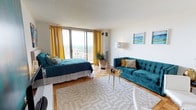 Located on the 13th floor at 4710 Bethesda Avenue, this thoughtfully designed studio offers an open, single-level layout that maximizes both function and style. The main living area is bright and welcoming, bathed in natural light from large floor-to-ceiling sliding glass doors that lead to a private balcony with sweeping views of the Bethesda skyline. Warm parquet hardwood flooring runs throughout, complementing the space's vibrant teal and gold color palette. A tufted teal velvet sofa anchors the living area, while a striking teal-painted accent wall frames the entertainment center, adding a bold focal point to the open-plan layout. The sleeping area flows seamlessly from the living space, offering comfort and versatility. The galley-style kitchen features white cabinetry, ample counter space, a gas range, dishwasher, and a peninsula with bar seating — ideal for casual dining. A breakfast bar with modern stools adds to the functional design. The full bathroom is clean and well-appointed, featuring a wide vanity with a mirrored medicine cabinet, tile flooring, and a shower/tub combination. Throughout the studio, curated artwork and lush greenery enhance the vibrant, elevated ambiance of this high-rise Bethesda residence.