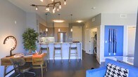 This single-floor space showcases a thoughtfully designed open-concept layout that seamlessly integrates the living and kitchen areas. The kitchen is a standout feature, centered around a marble-topped island with bar seating accommodating three gold-toned stools. Dark-finish cabinetry lines the walls, complemented by stainless steel appliances, including a full-size refrigerator and built-in microwave. Above the island, a series of pendant lights and a striking industrial-style pipe chandelier with exposed Edison bulbs provide both task and ambient lighting, adding a modern-industrial character to the space. Dark hardwood flooring runs throughout, creating a sleek, cohesive look that contrasts with the light gray walls. A dedicated in-unit laundry closet is conveniently tucked near the entry, housing a full-size stacked washer and dryer. The living area incorporates a warm wooden desk and lush greenery, lending an inviting, lived-in feel. A vibrant blue accent wall and matching blue armchair introduce bold pops of color, balancing the otherwise neutral palette. The overall ambiance is contemporary and functional.