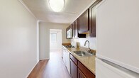This single-floor, 2-bedroom, 2-bathroom residence offers a functional and comfortable layout designed to maximize everyday living. The galley-style kitchen is a standout feature, equipped with rich dark-stained wood cabinetry, granite-toned countertops, a stainless steel sink, gas range, built-in microwave, and white appliances. Warm hardwood flooring runs through the kitchen and hallway, creating a clean visual flow throughout the common areas. The spacious primary bedroom is notably generous in size, carpeted for comfort, and includes a dedicated walk-in closet with built-in shelving on multiple walls, offering excellent organization and storage capacity. The second bedroom is also carpeted and features a window that brings in natural light. Both bathrooms are finished with white tile surround tub-shower combos; one features a dark wood vanity with a white countertop, a round mirror, and a mosaic-tile floor, while the second maintains a clean, bright aesthetic. Textured ceilings are present throughout, and crown molding accents the kitchen and hallway areas. The overall ambiance is neutral and move-in ready, with a light color palette of whites and warm beiges providing a calm, versatile backdrop throughout the entire residence.