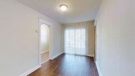 This single-floor residence offers a clean, functional layout with a warm and understated aesthetic throughout. The main living area is grounded by rich, dark hardwood flooring that contrasts beautifully with the light-toned walls, creating a welcoming and cohesive visual balance. A large sliding glass door dressed with vertical blinds serves as a prominent feature, allowing abundant natural light to flow into the space while providing access to the outdoors. The flush-mount ceiling light adds practical illumination, complementing the brightness brought in by the glass door. An elegantly arched doorway connects the main room to an adjacent area, which features carpeted flooring — offering a subtle transition in texture and tone between spaces. White baseboards and trim throughout the residence lend a crisp, finished look to each room. The overall ambiance is calm and neutral, providing a versatile canvas well-suited for a variety of interior styles. The thoughtful architectural details, including the arched passage and the interplay of flooring materials, add character to what is otherwise a straightforward and practical single-story layout.