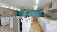 This dedicated laundry facility is designed with practicality and efficiency in mind, offering a clean and organized environment for shared use. The room is immediately identifiable by its bold teal accent wall, marked with the word "LAUNDRY" in prominent block lettering, providing a cheerful pop of color against the neutral grey wainscoting and white ceiling. A ceiling fan is centrally positioned to maintain comfortable air circulation throughout the space. Rows of white top-load washing machines are neatly arranged along the center and walls of the room, accompanied by a matching set of dryers lining the perimeter. The machines are organized in a functional layout that maximizes capacity while maintaining clear pathways for easy access. White cabinetry and partition panels between machine groupings provide a tidy, structured appearance. The floor is finished with durable, easy-to-clean tan tile, well-suited for a high-traffic utility space. Overhead fluorescent strip lighting ensures the entire room is brightly and evenly illuminated. Overall, the facility presents a well-maintained, organized, and highly functional space built to accommodate the laundry needs of a shared residential or commercial community.