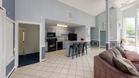Located at 5708 Trailview Ct in Frederick, MD, this single-floor community clubhouse offers a thoughtfully designed gathering space finished in a cohesive blue-gray and white color palette. The interior is unified by light ceramic tile flooring and classic wainscoting with white-trimmed paneling that lines the lower walls throughout, lending the space a polished, coordinated aesthetic.

The main lounge area is generously proportioned, featuring vaulted ceilings that enhance the sense of openness. Comfortable seating, including plush sofas arranged around an area rug, creates an inviting environment for relaxation or socializing. Multiple windows dressed with sheer white curtains allow natural light to fill the room, offering views of the surrounding greenery. A wall-mounted flat-screen television and a dark wood console table serve as focal points in the entry area.

The kitchen is fully equipped with gray cabinetry, granite-style countertops, black appliances including a range, microwave, and refrigerator, and a central island with bar seating for multiple guests — ideal for catered events or community gatherings. An adjacent utility room provides additional functional space.

Throughout the clubhouse, carefully chosen decorative accents, including framed artwork and a Village Development Group branded display, reinforce the professional, community-oriented character of this well-maintained facility.