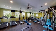 This fitness center in Conroe, Texas offers a comprehensive workout environment designed to support a wide range of exercise routines. The space is outfitted with bold yellow-green walls, carpeted flooring, and recessed lighting complemented by ceiling fans that keep the environment comfortable during workouts. Large windows along the perimeter flood the space with natural light and provide views of surrounding greenery, creating an energizing atmosphere.

The cardio zone features multiple treadmills, elliptical machines, and a stationary bike, all arranged along a full-length wall mirror that visually expands the room. A dedicated strength training area includes a multi-station cable machine and adjustable benches for a variety of resistance exercises. A dumbbell rack stocked with a full range of free weights provides additional options for strength training.

A functional training corner adds versatility to the facility, housing a multi-purpose rig with pull-up bars and suspension straps, as well as a heavy-duty Everlast punching bag for combat-style conditioning. Wall-mounted televisions are positioned throughout for entertainment during workouts. The center also includes a water station and a dedicated restroom for convenience. Posted fitness center rules ensure a safe and organized environment for all users.