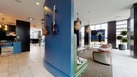 The lobby at 255 Huguenot Street in New Rochelle, NY makes a striking first impression with its bold, music-inspired design and carefully curated mix of materials and textures. The reception area features reclaimed wood wall panels, dark subway tile accents, and a patterned tile backsplash, creating a warm yet industrial aesthetic. Blue accent surfaces and industrial metal railings add modern contrast throughout.

At the heart of the lobby is a distinctive lounge area anchored by a sweeping tufted green leather banquette set against a vibrant cobalt blue curved wall, adorned with a collection of electric guitars as wall art. Pendant chandeliers and a sputnik-style light fixture provide ambient illumination, while tan leather armchairs and mixed side tables complete the seating arrangement.

Floor-to-ceiling windows along the perimeter flood the space with natural light and offer open views of the surrounding streetscape and landscaping. Light stone tile flooring runs throughout, providing a neutral foundation that complements the bold décor. A cozy private dining or conference area is accessible via an arched corridor, furnished with a large dark dining table, leather cantilever chairs, and a candle-style chandelier. Tall indoor plants punctuate the space, adding organic warmth to this dynamic and welcoming lobby.