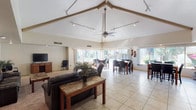 This single-floor space is defined by its dramatic vaulted ceilings with exposed beam accents, ceiling fan, and a track lighting system that runs the full length of the open-plan interior. Light-toned tile flooring extends throughout, complemented by a carpeted lounge area that adds warmth and definition to the seating zone.

The spacious lounge area is furnished with dark leather sofas and armchairs arranged around a marble-top coffee table, with a wall-mounted mirror and flat-screen television on a wooden media console adding functional detail. Recessed lighting and large windows provide excellent illumination throughout this area.

Adjacent to the lounge, a generous dining area features multiple high-top tables with dark upholstered chairs, all positioned to take full advantage of the expansive floor-to-ceiling sliding glass doors and large windows. These openings frame views of landscaped outdoor greenery and flowering shrubs, allowing natural light to flood the interior.

A kitchen bar area with a dark countertop and modern bar stools offers a casual seating option, with a pass-through view into a kitchen space beyond. Additional windows along this wall maintain the bright, airy character consistent throughout the space.

Overall, the interior presents a clean, neutral palette, with high ceilings and generous glazing creating an open, welcoming ambiance suitable for communal gathering and multi-purpose use.
