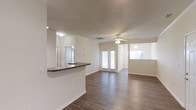 This single-floor space offers an open and airy layout designed for comfortable everyday living. The main living area features rich wood-look hardwood flooring that flows seamlessly throughout, lending warmth and continuity to the space. A ceiling fan with a light fixture keeps the area well-ventilated and well-lit, while large windows and a set of classic French doors flood the interior with abundant natural light, creating a bright and welcoming atmosphere.

The kitchen is positioned adjacent to the living area and is defined by a raised bar-height counter finished with a dark granite-style surface, providing a practical and stylish transition between the two spaces. A mirror mounted near the kitchen enhances the sense of openness. The kitchen pass-through design encourages an inclusive, social environment ideal for entertaining or everyday interaction.

The walls are finished in a clean, neutral tone that complements the flooring and contributes to the overall light and open feel of the space. Visible HVAC vents in the ceiling indicate a central air system for year-round climate control. The French doors lead to what appears to be an outdoor area, offering the potential for seamless indoor-outdoor living. Overall, this space balances functional design with a clean, modern aesthetic.