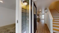 This two-story townhouse in Niantic, Connecticut offers a practical and well-appointed layout across two floors. The entry level introduces a covered entryway with a nautical-style wall sconce and lap-siding detail, lending a coastal character to the arrival experience. A dark-framed barn-style door leads into the main living area, where light wood-style flooring extends throughout, complementing the clean white walls and creating a bright, open atmosphere.

The kitchen is equipped with gray cabinetry, ample counter space, a full suite of white appliances including an electric range with overhead microwave, dishwasher, and sink. The adjacent living area is generously proportioned with recessed lighting and a wall-mounted mini-split system for climate control. A half bath is conveniently located on this level.

The upper floor houses three bedrooms, each featuring wood-style flooring and windows that allow natural light and views of the surrounding greenery. The primary bathroom includes a double-sink vanity with a white countertop and a walk-in shower, while the second full bathroom offers a single vanity and a shower-tub combination. An attached one-car garage with automatic opener and a paved driveway rounds out this townhouse's functional amenities.
