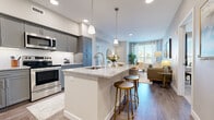 This thoughtfully designed single-floor residence offers a cohesive blend of modern finishes and comfortable living spaces. The open-concept layout connects the kitchen, dining, and living areas seamlessly, creating an inviting and functional environment. The kitchen is a standout feature, equipped with stainless steel appliances including a refrigerator, range, and microwave. Gray shaker-style cabinetry pairs elegantly with granite countertops and a white subway tile backsplash. A central island with a built-in sink and bar seating serves as both a prep space and a casual gathering spot, illuminated by pendant lighting above. The living room is bright and airy, benefiting from large windows that allow ample natural light to fill the space, complemented by wood-style flooring throughout the main areas. A secondary living or flex room features soft carpet, a soothing accent wall, and additional window views. The bedroom is a serene retreat with plush carpet, natural light from a large window, and access to a spacious walk-in closet. The bathroom mirrors the home's refined aesthetic with granite-topped gray vanity cabinetry and wood-look tile flooring. The entryway is welcoming and practical, leading smoothly into the heart of the home.