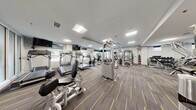 This fitness center is thoughtfully designed across a single level, comprising two distinct areas that together provide a comprehensive workout environment. The main gym area features patterned commercial-grade carpet with yellow accent striping and is illuminated by recessed panel lighting throughout. Large windows along one wall allow natural light to filter in and provide views of the surrounding greenery, creating an open and energizing atmosphere. This primary space is extensively equipped with Hoist-brand cable machines, multi-station strength systems, a full dumbbell rack, and a row of cardio machines including treadmills, ellipticals, and stationary bikes. Wall-mounted televisions are positioned near the cardio equipment for entertainment during workouts. Floor-to-ceiling mirrors line key sections of the gym, enhancing the sense of space and allowing users to monitor their form. The second room serves as a dedicated studio space, finished with wide-plank wood-look flooring in a neutral gray tone. This area is outfitted with a wall-length mirror, a ballet barre, wall-mounted exercise mats, and stability balls, making it well-suited for yoga, barre, stretching, or group fitness classes. A wall-mounted monitor and exterior-facing windows with roller shades complete the studio. Together, both spaces offer a clean, modern, and fully functional fitness facility.