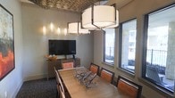 This sophisticated single-floor meeting and dining space combines functional design with refined aesthetic details. At its centerpiece is a large rectangular wooden conference table surrounded by cushioned leather-accented chairs, accommodating a generous number of guests. Overhead, a striking cluster of drum-style pendant chandeliers with white shades and dark metal frames commands attention, while a decoratively textured ceiling panel adds an additional layer of architectural interest.

The walls feature a subtle, textured neutral finish that lends warmth to the room, complemented by a series of slim vertical accent lights that glow softly along one wall. A wooden media console sits against the back wall, housing a flat-screen television alongside a telephone and decorative accessories, making the room well-suited for both presentations and private gatherings.

Large windows along the adjacent wall flood the space with natural light and offer a clear view of an outdoor courtyard, creating a pleasant connection to the exterior environment. A sculptural branch-like centerpiece rests on the table, adding an organic and artistic touch. The overall ambiance is polished yet welcoming, blending contemporary furnishings with thoughtful design elements to create a versatile and visually engaging space.