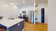 This single-floor residence is designed with a clean, modern aesthetic that emphasizes an open and functional layout. The kitchen serves as a central feature, anchoring the living space with a navy blue island topped with white quartz countertops and an integrated sink. Pendant globe lights hang above, adding a contemporary touch. The kitchen is fully equipped with stainless steel appliances, including a refrigerator, electric range, and microwave, complemented by two-tone cabinetry — white upper cabinets paired with navy lower cabinets. Open wood shelving near the entry adds both storage and visual interest.

The adjoining dining and living areas share an open, connected feel, with warm hardwood-style flooring running throughout. The living room features a large sliding window that brings in natural light and offers views of the surrounding neighborhood. Recessed lighting illuminates both the kitchen and dining areas effectively.

The bedroom is a calm retreat, equipped with a wall-mounted split AC unit and direct access to a spacious walk-in closet with wire shelving and built-in dresser space. The bathroom features light tile flooring, a warm wood vanity, and a clean, minimal design. Overall, the space maintains a cohesive, contemporary character.