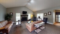 Located at 3259 Alden Pond Ln in Eagan, Minnesota, this single-floor community space offers a thoughtfully designed layout that balances comfort and functionality. The centerpiece of the space is a generously sized living area highlighted by soaring vaulted ceilings and a striking stone-surround fireplace with a wood mantel, above which a flat-screen television is mounted. Multiple windows dressed with classic shutters and blinds flood the room with natural light and offer views of the surrounding greenery. The living area is furnished with leather sofas and upholstered wingback chairs arranged to encourage conversation and relaxation. An adjacent dining area features dark wood tables and chairs accented by a trio of decorative octagonal mirrors on the wall, adding visual interest to the space. The fully equipped kitchen is outfitted with rich dark cabinetry, granite countertops, hardwood flooring, stainless steel appliances including a refrigerator, range, and microwave, and a deep double-basin sink beneath a pendant light. A welcoming entryway with a cushioned wooden bench, a large framed mirror, and direct access to the outdoors completes the space. Throughout, a consistent warm gray palette and quality finishes contribute to a cohesive, inviting ambiance.
