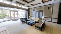 This refined lobby space makes a strong architectural statement with its striking coffered ceiling, finished in a rich dark wood trim that contrasts elegantly against the cream-toned panels and recessed lighting. The design carries a formal, upscale character throughout, with carefully framed wall panels and a neutral color palette that lends a sense of warmth and polish.

The seating area is thoughtfully arranged, featuring a cream upholstered sofa, patterned accent chairs, and sleek dark-framed benches centered around a low-profile coffee table. Decorative table lamps add ambient lighting and a refined touch to the arrangement. The space is grounded by a large striped area rug that defines the lounge area within the broader open floor plan.

Natural light floods the space from a glass-paneled entryway at the front, offering views to the exterior and enhancing the airy, open feel of the interior. Elegant curtain panels frame the entry corridor, adding texture and a soft architectural detail. Interior doors with frosted glass panels provide access to adjoining areas while maintaining a cohesive, sophisticated aesthetic. Overall, this lobby exudes a professional and welcoming ambiance, ideal for making a lasting first impression.