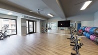 This well-appointed fitness studio offers a clean, contemporary design ideal for a variety of group and individual workouts. The space is finished with warm hardwood-style flooring throughout, providing a durable yet inviting surface suited for cardio, stretching, and functional training activities. Large windows along one wall flood the interior with natural light, while a ceiling fan ensures comfortable air circulation during intense sessions. A row of stationary cycling bikes lines one side of the studio, complemented by a well-stocked medicine ball rack offering a range of weights for strength and conditioning exercises. Colorful step platforms provide versatility for aerobic and interval training routines. Full-length wall mirrors expand the sense of space and allow users to monitor their form during workouts. A large flat-screen television is mounted prominently on the mirrored wall, supporting instructor-led or on-demand fitness classes. The studio also features an adjacent secondary area with additional equipment, including stability balls and a folding mat station, connected via an interior pass-through window that maintains an open, connected feel. Access to an outdoor balcony is available through glass-paneled double doors, offering a refreshing connection to the outdoors. The overall ambiance is bright, spacious, and thoughtfully organized to support a comprehensive fitness experience.